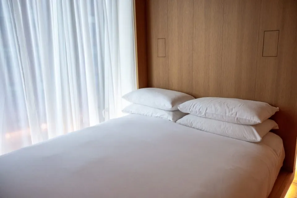 A double bed with white bedlinen and stacked pillows, against a wood-panelled wall beside sheer curtains.