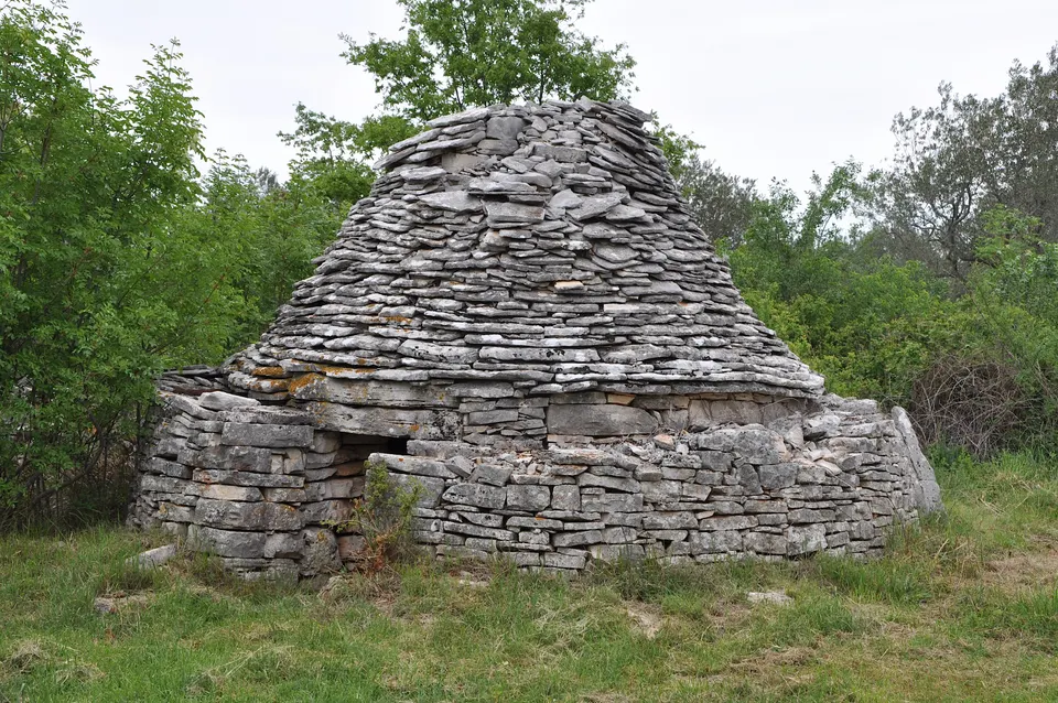 A kažun with a wide, low stone roof, surrounded by shrubs and grass.