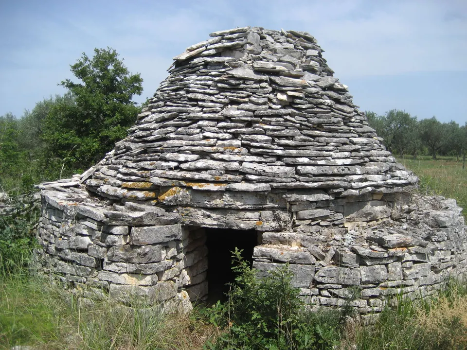 A broad kažun with a stepped stone roof, its low doorway framed by green shrubs.
