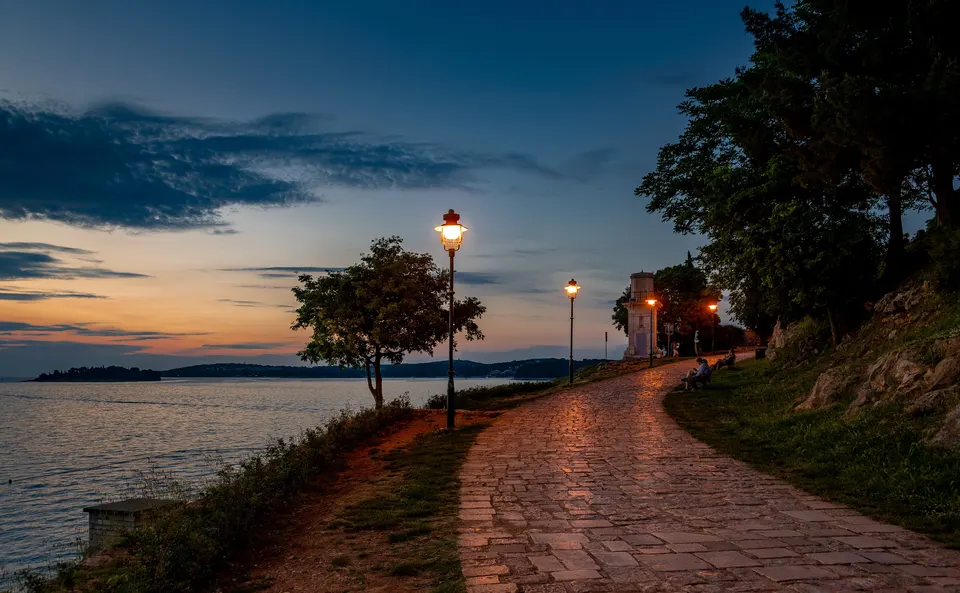Die Uferpromenade von Rovinj zur blauen Stunde, mit brennenden Laternen entlang des Steinwegs.