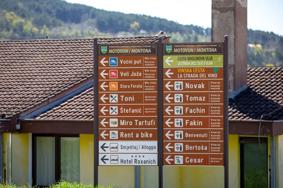 Un alto palo segnaletico in legno a Motovun indica produttori locali, un noleggio di biciclette e alloggi, davanti a un edificio con il tetto di coppi.