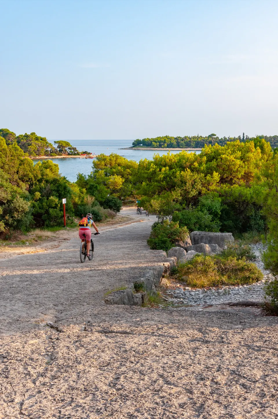 Ein Radfahrer auf einem sandigen Küstenweg zwischen niedrigen mediterranen Sträuchern, dahinter das Meer und eine bewaldete Insel.