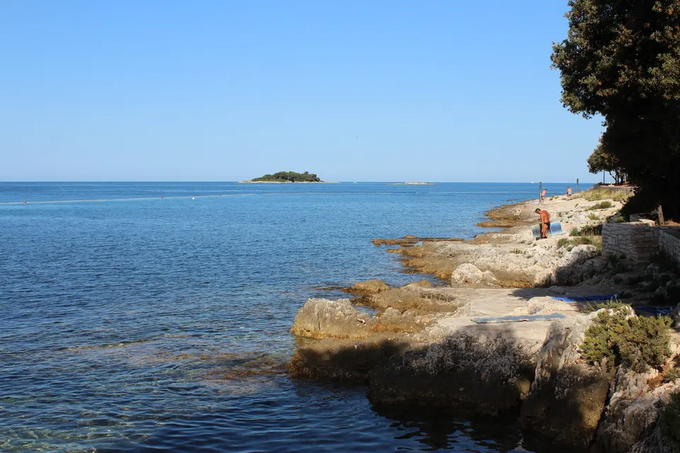 Flache Kalksteinfelsen und klares Wasser an der Küste eines Campingplatzes bei Vrsar (Orsera), vor der Küste eine kleine bewaldete Insel.