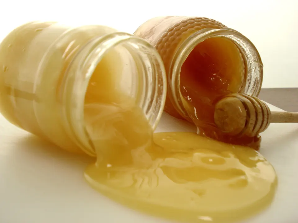 Due vasetti rovesciati di miele cristallizzato, uno chiaro e cremoso e uno più scuro, da cui cola miele denso su una superficie bianca accanto a un mestolino di legno.