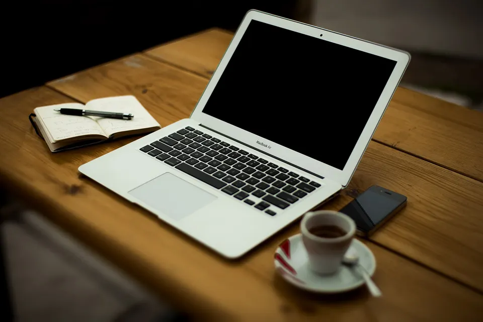 Ein aufgeklappter Laptop, ein Notizbuch mit Stift, ein Telefon und eine Espressotasse auf einem rustikalen Holzschreibtisch.
