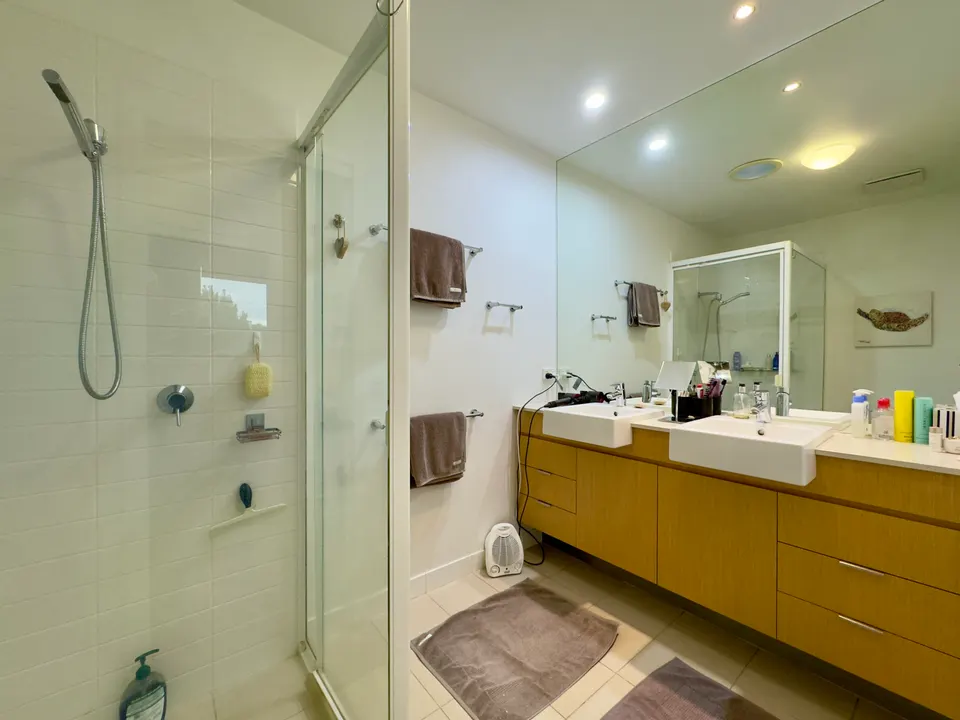 A glass shower beside a long vanity with drawers, two basins and a mirror that runs the full length of the counter, lit from above.