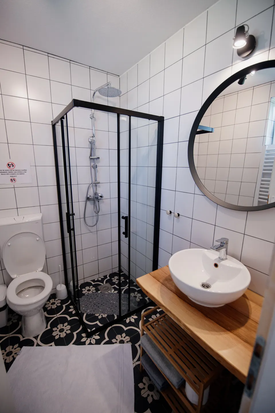 A small bathroom with a corner shower in black-framed glass, a round mirror, a white bowl basin on a timber vanity with an open shelf, and patterned floor tiles.