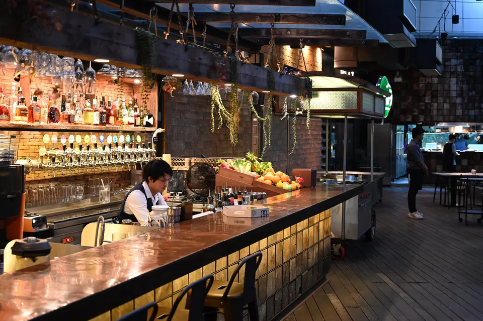 Eine lange Bar mit kupferfarbener Platte und sanft beleuchteter, gefliester Front, darüber hängende Gläser, Flaschen an der Rückwand und der Barkeeper bei der Arbeit hinter der Theke.