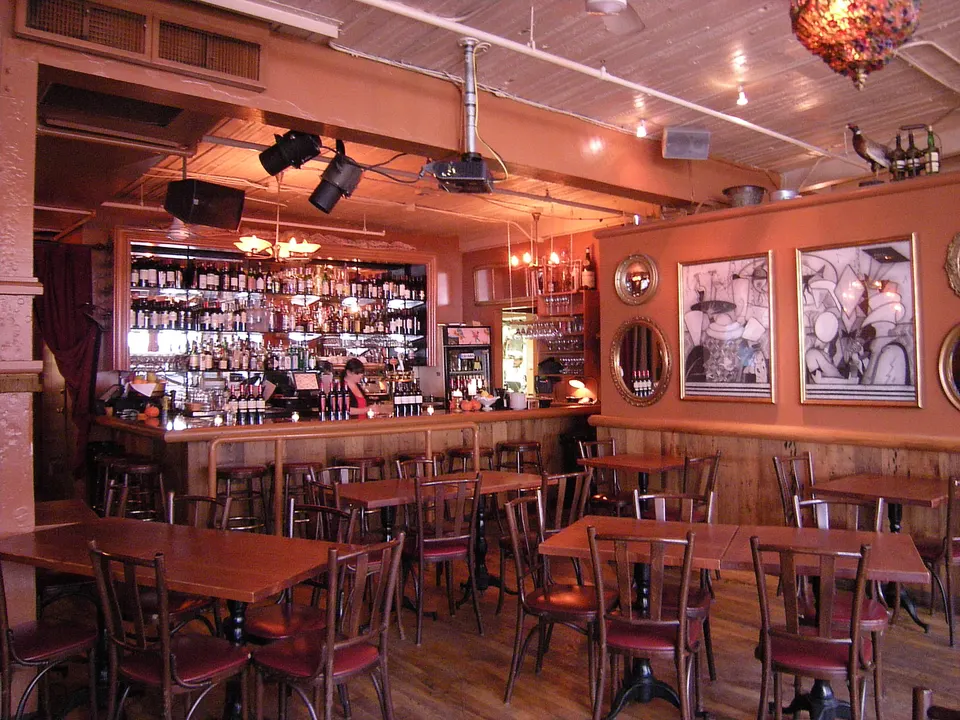 Eine Restaurantbar vom Gastraum aus gesehen: eine Holztheke mit Fußstütze und Hockern, ein beleuchtetes Rückbuffet mit Flaschen und gedeckte Tische ganz in der Nähe.