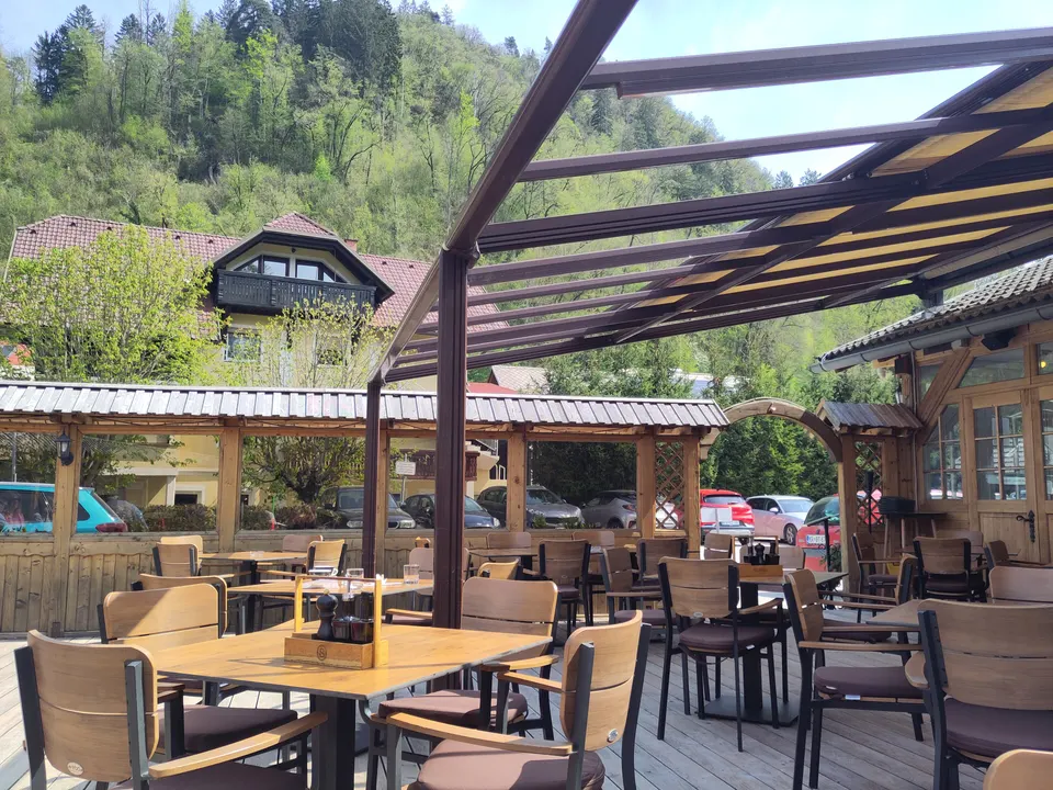 A restaurant terrace on a timber deck under a steel pergola with a translucent roof, set with wooden tables and woven chairs.
