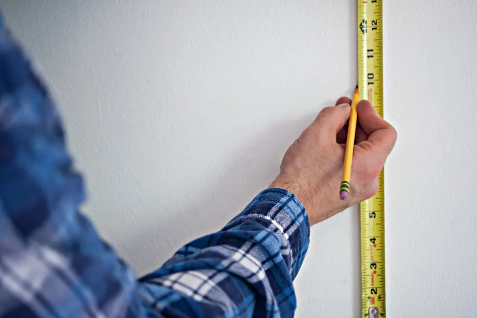 A tape measure held against a white wall while a pencil marks a height. Quantities in a good bill of quantities are measured, not guessed.