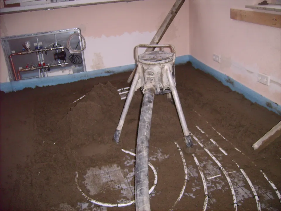 Screed being pumped over underfloor heating pipes, with the heating manifold on the wall. In a quote this is usually one line priced per m², and the description should say how thick it is.