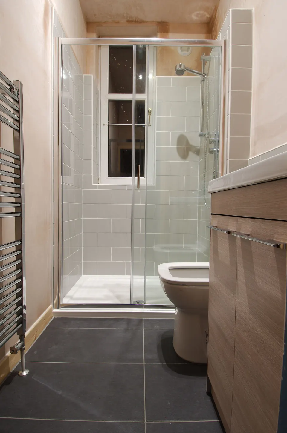 A finished narrow bathroom: a glass shower enclosure under the window, grey tiles, a wood-effect vanity unit and a heated towel rail.