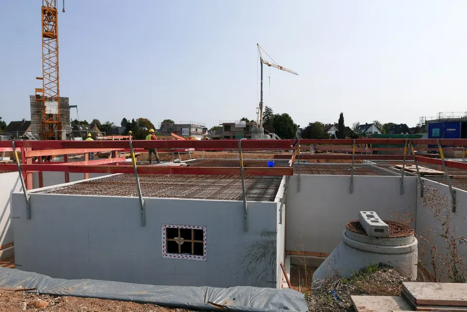 A house shell on a building site: grey block walls, steel reinforcement on the slab, red edge protection and tower cranes behind.