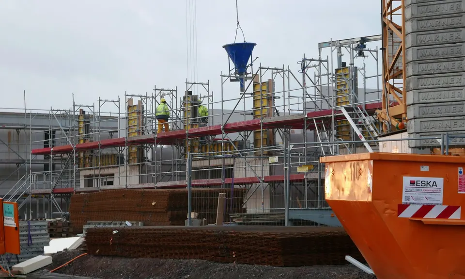 Eine Baustelle mit Gerüst, einem Betonkübel am Kran in der Luft und einem orangefarbenen Container im Vordergrund, jener Art Behälter, auf die sich die Schuttabfuhr (odvoz šute) bezieht.
