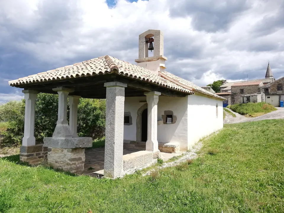 The Chapel of St Roch in Draguć: a small white church with a stone bell gable and a tiled porch on stone columns, on a grassy slope.