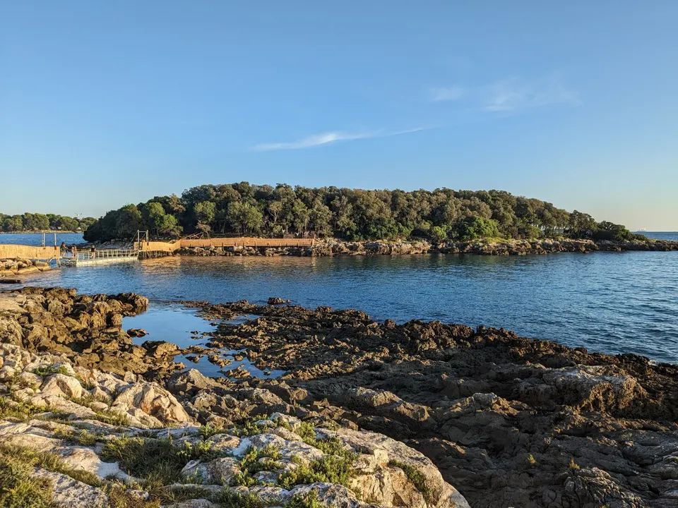 The wooded islet of Školjić off Funtana in late-afternoon light, with a walkway to the shore and dark rocks in the foreground.