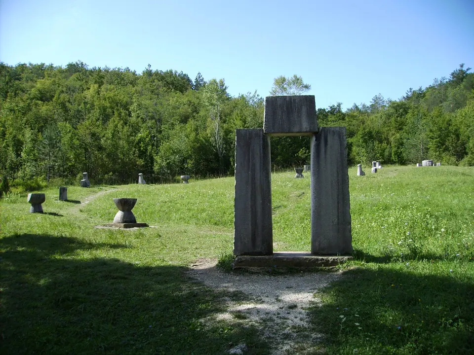 One of the monuments of the Glagolitic Alley near Hum: a tall stone gate on a grassy meadow, with smaller stone seats scattered around it and forest behind.