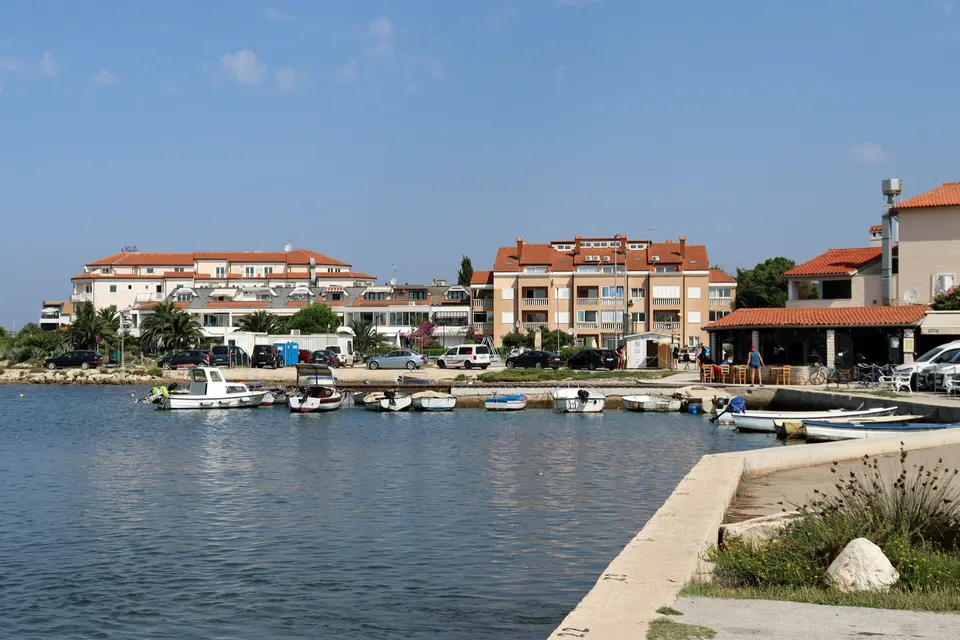 Small boats moored in a calm corner of Medulin Bay, with apartment buildings and palm trees along the waterfront.