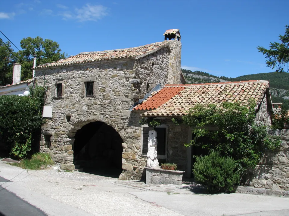 The north gate of Roč: a rounded stone archway beneath an old house with a tiled roof, beside a small garden and a white stone sculpture.
