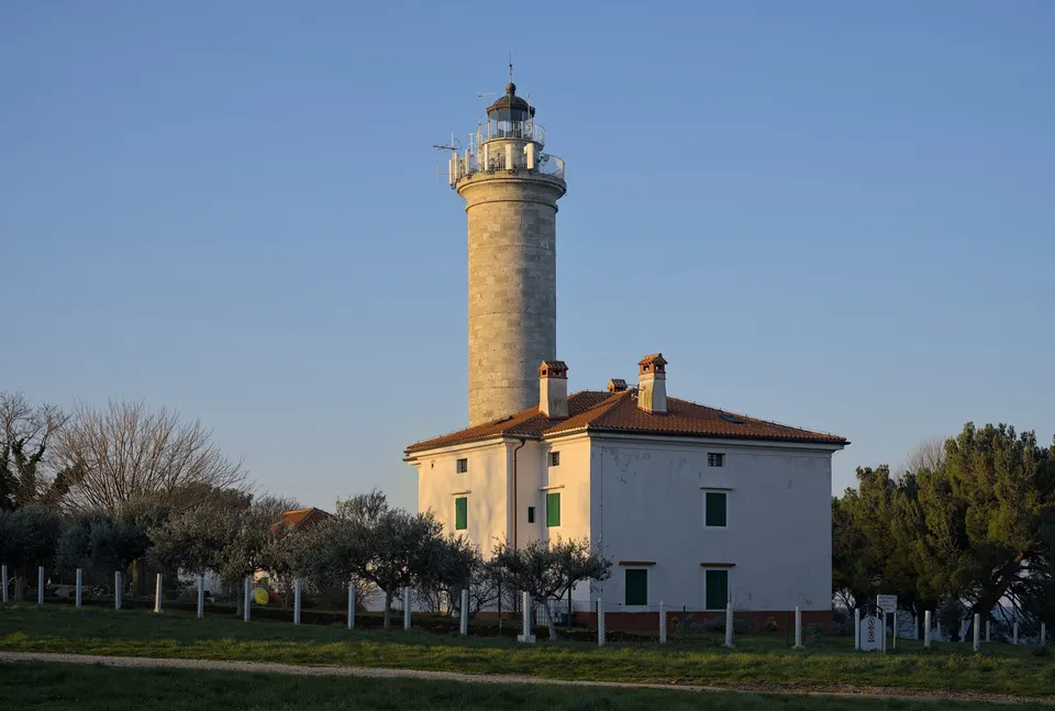 Savudrija lighthouse in the evening light: a tall stone tower rising from the white keeper's house, among olive trees and pines.