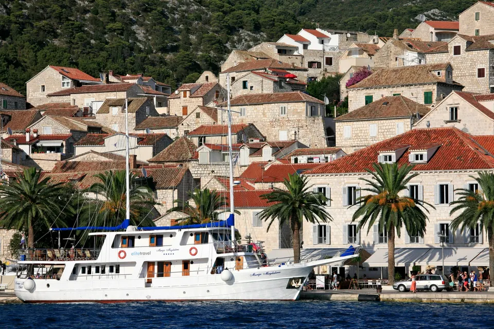 Stone houses of Hvar town climbing the hillside above the harbour, with palm trees and a white excursion boat on the waterfront.