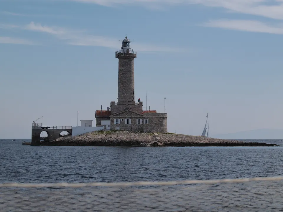 The stone tower and buildings of Porer lighthouse on its tiny, flat islet, with a sailing boat passing behind.