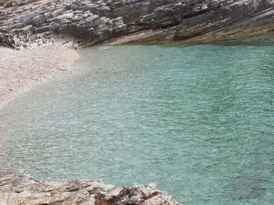 A small cove at Kamenjak, where layered rock gives way to a curve of pale, fine pebbles and very clear water.