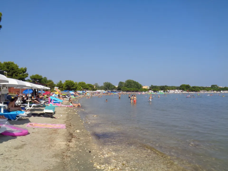 The shallow, sandy edge of the bay at Medulin, with swimmers wading far out in calm water.
