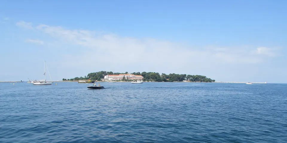 Sveti Nikola island, low and wooded, seen across the water from Poreč with boats moored offshore.