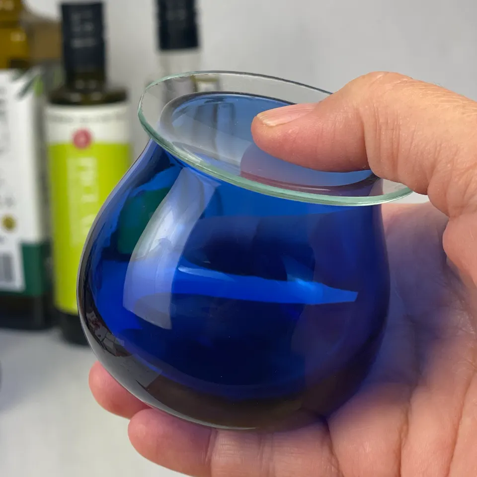 A professional olive oil tasting glass in cobalt blue, held in the hand with a glass lid on top.