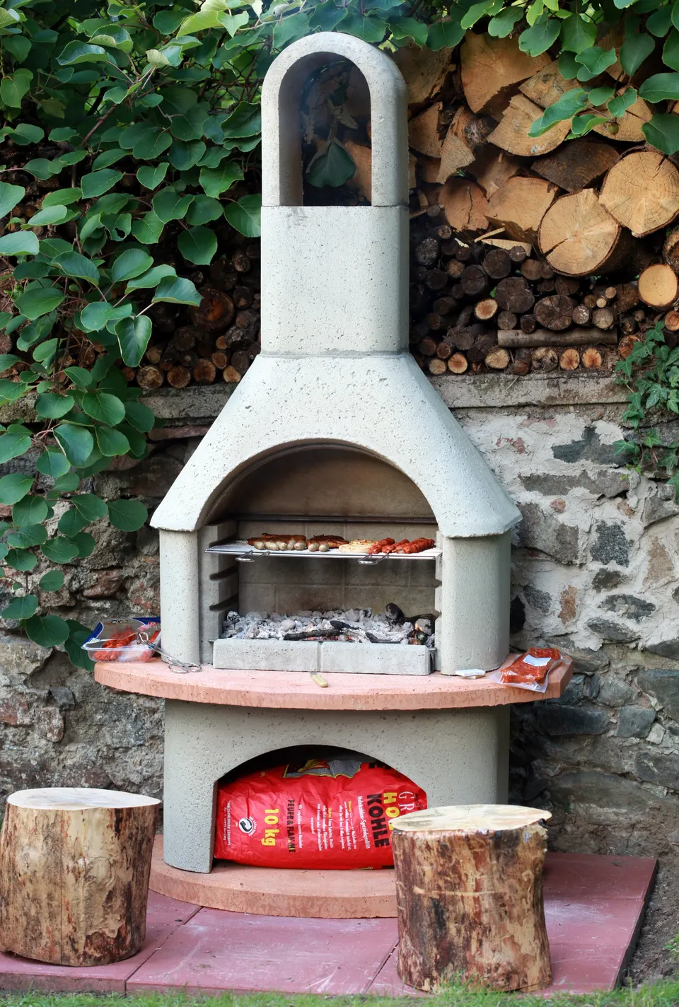 A masonry grill against an old stone wall, with firewood stacked behind it and log stools in front.