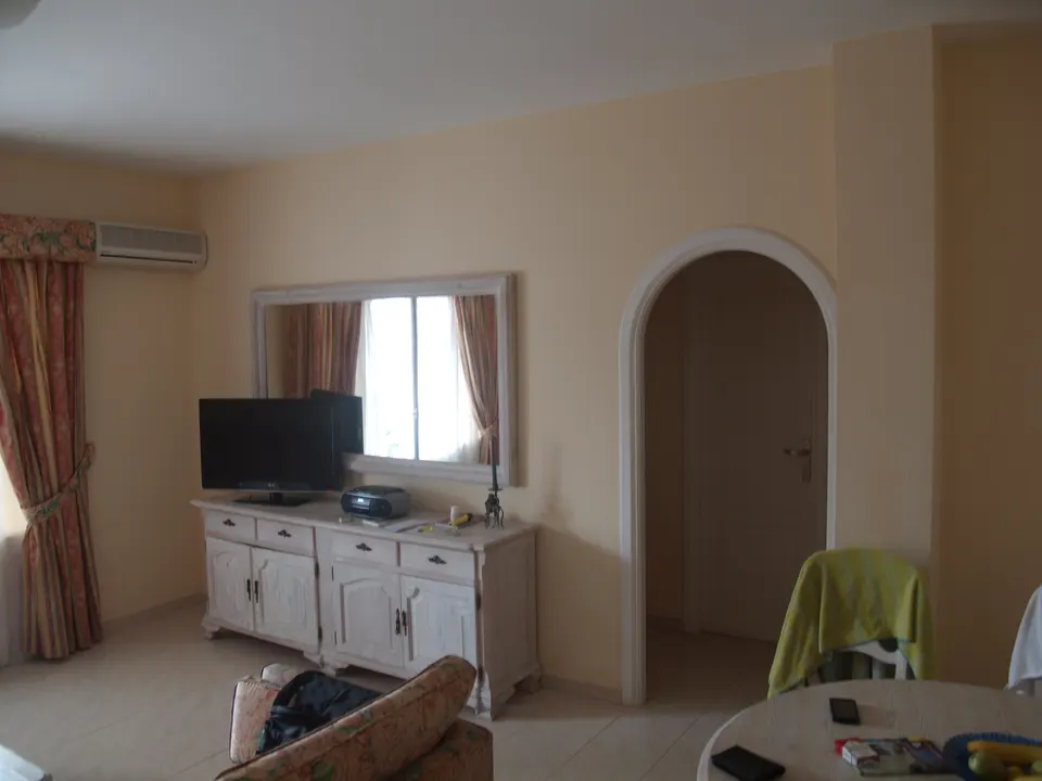 The living room of a holiday apartment, with a white sideboard and TV, a mirror, an arched doorway and a round table.