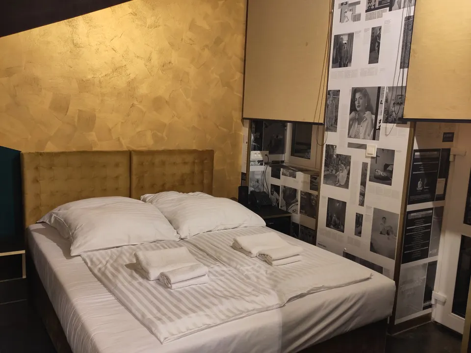 A hotel bed with a tall upholstered headboard, striped white linen and folded towels, beside a wall of framed newspaper prints.