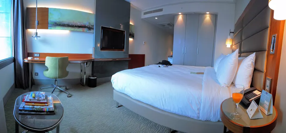 A modern hotel room with an upholstered headboard, a long desk with a chair, a wall-mounted TV and a round side table.