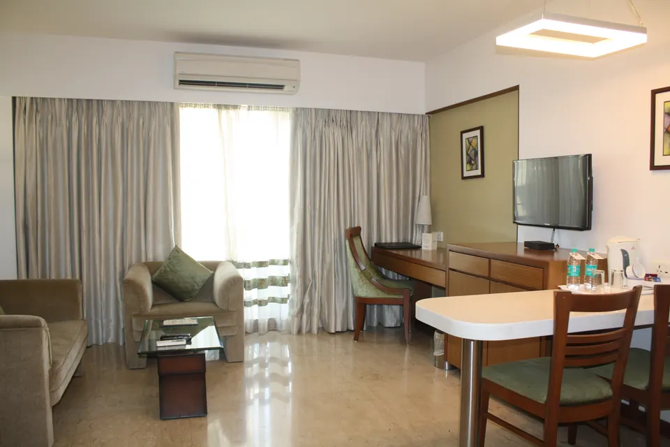 A serviced apartment with armchairs, a coffee table, a desk, a kitchen counter with stools and long curtains at the window.