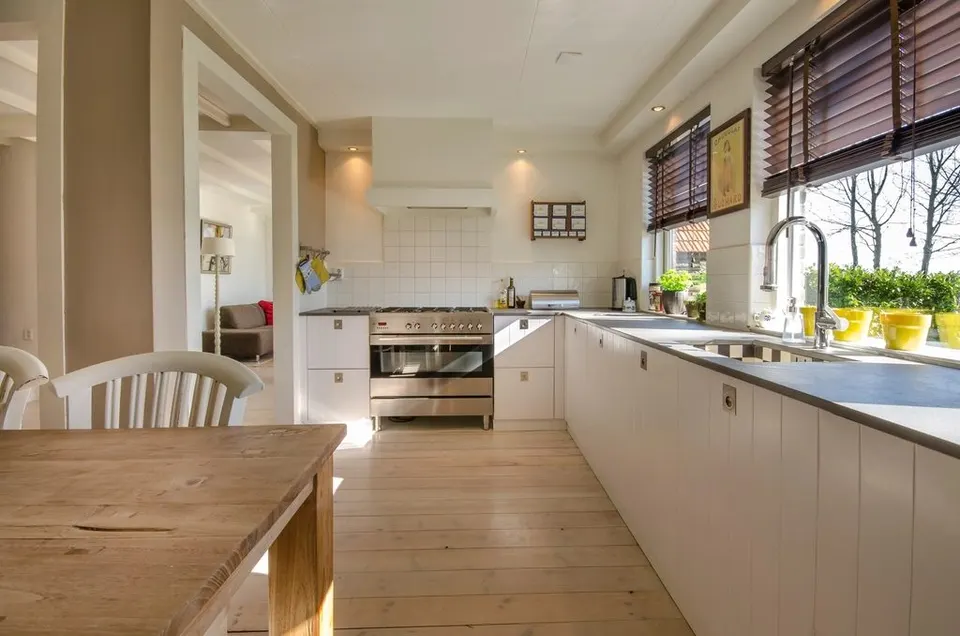 A long run of pale kitchen cabinets with a range cooker and a sink under the window, opposite a wooden dining table.